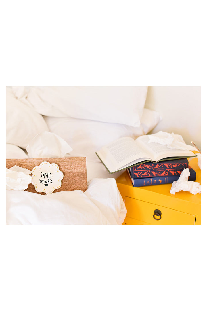 Happy Everything mini wood tissue box with DND mode tag on white bedding beside yellow nightstand with books