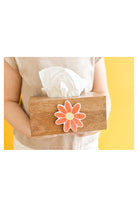 Hands holding a wooden tissue box with orange daisy flower decoration against yellow background
