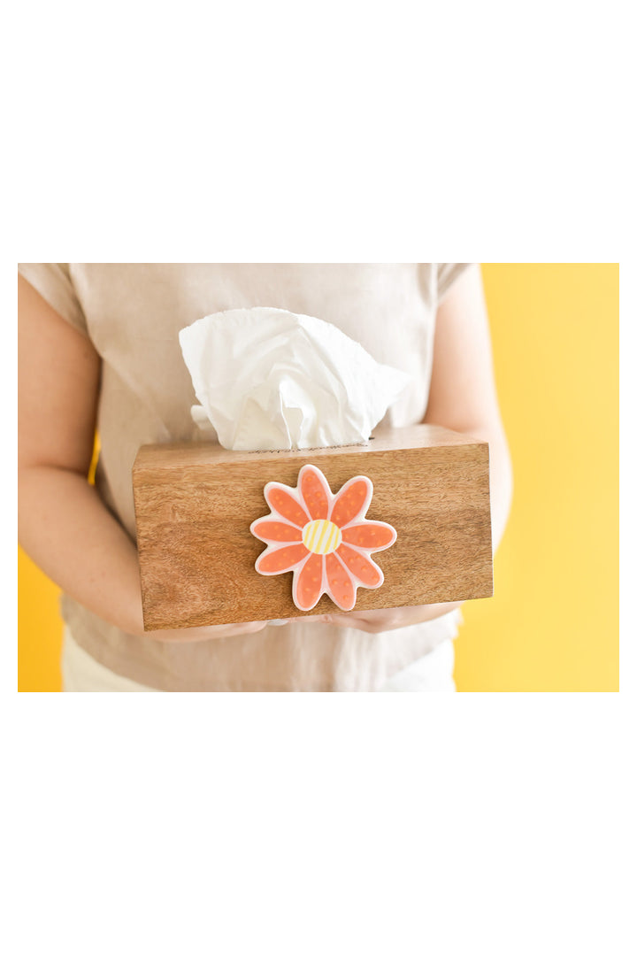 Hands holding a wooden tissue box with orange daisy flower decoration against yellow background